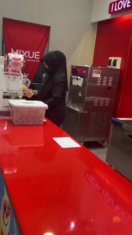 A person wearing a black hijab and a black t-shirt with writing on the back is working behind a red counter. They are holding a brown bottle and appear to be dispensing something into a plastic container filled with small cups. To the left of the person is a white ice cream machine with a red accent, and a red banner with the word "MIXUE" on it. To the right of the person is a silver ice cream machine. In the background, there is a stainless steel sink and two red dispensers with white labels. The environment appears to be an indoor food establishment.