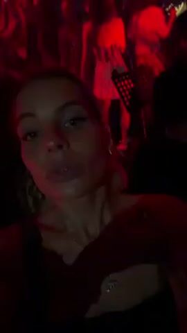 Two young women are posing in what appears to be a nightclub or similar venue. The lighting is predominantly red, casting a deep red hue over the entire scene. They are close together, with one person holding the camera and the other appearing to be standing behind or beside them. The camera angle is low, looking slightly upwards at the two individuals. The background is dark and blurry, suggesting other people and the general ambiance of a crowded space. They are both making different faces to the camera.