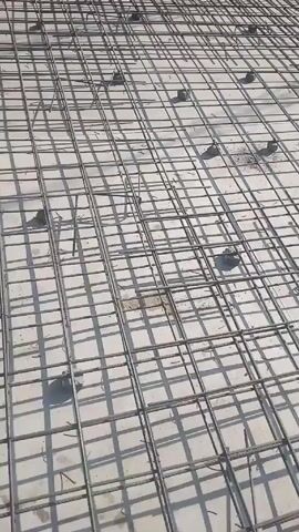 The scene showcases a construction site, possibly the foundation or floor of a building, where a grid of steel rebar is laid out. Small, dark cylindrical supports elevate the rebar from the concrete slab. In the background, completed buildings and surrounding vegetation are visible under a bright sky. The rebar grid suggests a structure designed to withstand heavy loads or stresses, typical of modern concrete construction.