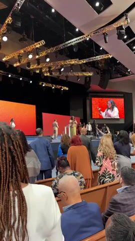 The video shows the interior of a large, modern church during a service. In the foreground, a diverse congregation is seated in wooden pews, facing a stage. The lighting is dramatic, with spotlights illuminating the stage and large screens displaying images. On stage, several individuals are visible. One woman, wearing a light-colored robe, stands at a podium and appears to be speaking, her image also displayed on a large screen to the right. Other individuals, including men and women in formal attire, are also on stage, some interacting with each other and with the congregation. The overall atmosphere suggests a lively and engaging religious service. The camera is positioned behind the congregation, looking towards the stage, providing a view of both the attendees and the performers.