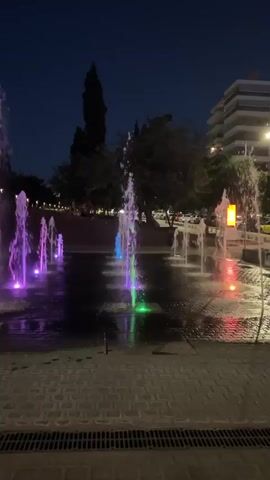 The video shows a nighttime view of a water fountain display. The fountains are lit with various colored lights, predominantly purple and green. The water shoots up in streams, creating a dynamic visual effect against the dark sky. Trees and buildings are visible in the background. The ground in the foreground is paved with interlocking bricks and has a grate. The overall atmosphere is serene and visually appealing, suggesting a public park or plaza designed for evening entertainment or ambiance.