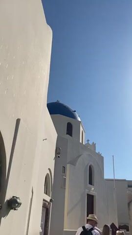 A vertical video begins from a low angle, looking up at a white building with a blue dome. The sky is a clear, bright blue with the sun shining from the right side of the frame. To the left, a large, white wall curves upwards, dominating the left side of the screen. To the right, a white church-like structure is visible, featuring arched windows and a prominent blue dome. Two red doors are present on the church facade. Two people are partially visible in the bottom right corner of the frame. One person wears a hat and a backpack. The video pans slightly upwards, offering a closer view of the church's dome and facade. The camera then appears to move, and the view shifts slightly, with the white wall on the left continuing to be prominent. The shot remains focused on the building and the sky.