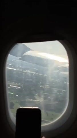 The video shows an airplane wing through an airplane window. The wing is mostly white and reflects sunlight. Below the wing, the ground is visible, appearing as a mix of gray and green, possibly showing trees and land features. A smartphone is visible at the bottom of the frame, apparently being used to record the view.