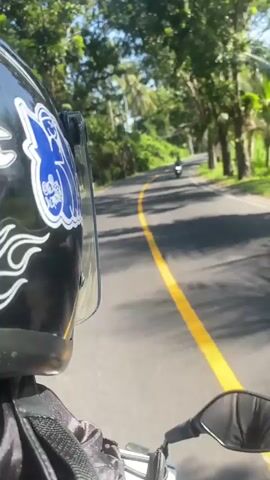 A first-person perspective video shows a person riding a motorcycle on a paved road lined with lush green trees and palm trees. The rider is wearing a black helmet with a blue and white design. A yellow line is visible on the road, dividing the lanes. In the distance, other motorcycles and vehicles can be seen. The sun is shining, casting shadows on the road. The overall scene suggests a tropical or subtropical environment.