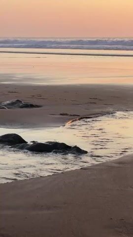A close-up, low-angle shot shows a stream of water flowing over sand on a beach towards the ocean. The water is reflecting the sunset sky. The sky is a pale orange and pink. The ocean has gentle waves. The sand is wet in places and dry in others. There are a few dark rocks visible in the water. The video is calm and serene.