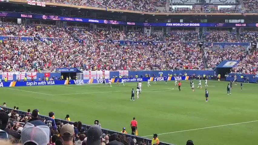 The video captures a wide shot of a soccer match taking place in a large, packed stadium. The field is a vibrant green, with white markings indicating the boundaries and penalty areas. Players in white and dark blue uniforms are scattered across the field, engaged in play. The crowd in the stands is a blur of colors, with many spectators visible, suggesting a high-turnout event. Several large screens are mounted above the stands, displaying text that appears to be related to the event, including "FIFA WORLD CUP 2026" and "MIAMI". Banners, some with national flags, are also visible along the sidelines. The perspective is from a seated spectator in the lower level of the stadium, looking towards the field. The overall atmosphere appears to be energetic and exciting, typical of a professional sporting event.