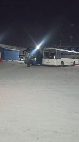 At night, a white bus is positioned in a snow-covered lot. The bus drives backward out of its parking spot, producing a cloud of exhaust. A person in a neon yellow reflective vest stands near the back of the bus. There are two other buses, one white and one blue, parked in the lot. A prefabricated building sits in the background, with illuminated windows.  A tall antenna can be seen in the distance.