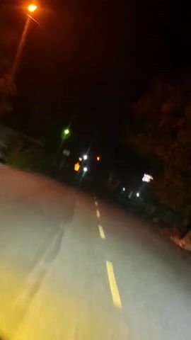 The video is a first-person perspective of a vehicle driving at night on a paved road. The road has a yellow dashed line marking the center. The driver's headlights illuminate the road ahead and create a bright yellow-green hue. In the distance, other vehicles are visible with their taillights illuminated. Streetlights and other light sources are also present along the side of the road, which is lined with vegetation and structures. The video is shaky, suggesting movement and potentially a bumpy ride. The overall impression is of a car journey through a dimly lit rural or suburban area at night.