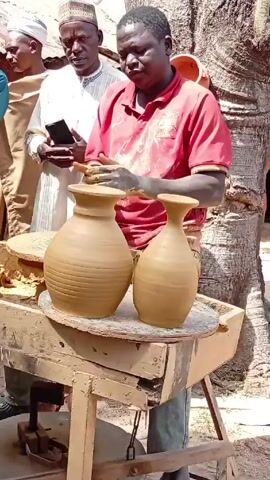 A man in a red shirt stands at a pottery wheel outdoors, working on two clay vases. He uses a small tool to refine the shape of one of the vases. Three men stand behind him, observing. One of the men is holding a smartphone. The wheel is wooden and appears to be manually operated. The vases are a light brown color and have a distinct shape with a narrow neck. A bucket of clay and other clay materials are visible on the wheel's surface. The background shows a building and some trees, suggesting a village or rural setting.