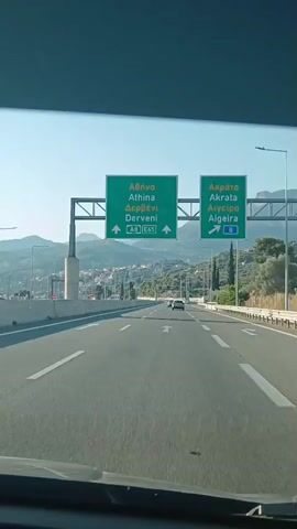 The video shows a view from a car driving on a highway. The road has multiple lanes and white markings. In the distance, there are mountains and a town with buildings. Overhead signs indicate directions to Athens, Derveni, Akrata, and Aigeira, with route numbers A8 and E45 visible. A few other cars are on the road ahead. The sky is clear and blue.