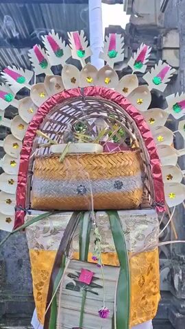 The video shows a close-up of an elaborately decorated offering or altar piece, likely in a Balinese Hindu temple setting. The centerpiece is a woven basket containing offerings, possibly food and flowers, enclosed within a semi-circular structure. This structure is adorned with layers of woven palm leaves, some of which are painted with pink and green accents, and topped with a red woven border. Below the basket, a tall, cylindrical object is wrapped in fabric. The fabric appears to be golden with intricate patterns, and it's decorated with green palm leaves and more woven leaf elements. The background suggests an outdoor environment with stone architecture and an overcast sky. The camera slowly zooms in on the offering. There are no visible people in the frame.