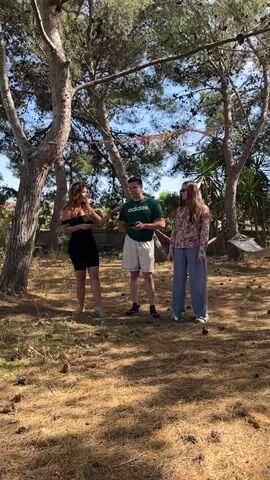 Three people are standing outdoors on a sunny day. Two women and one man are standing in a triangle formation on a surface covered with fallen pine needles and pine cones. The woman on the left wears a black outfit, the man in the center wears a green T-shirt, khaki shorts, and is looking down at a smartphone. The woman on the right wears a patterned floral shirt and light blue pants. Behind the people are several trees, and in the background there is a hammock strung between two trees. The woman on the left holds a yellow object and the woman on the right holds a pink and white object in her hand.