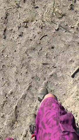 A first-person perspective video shows a person walking on a dirt path. The person is wearing purple pants and hiking boots. The path is covered in dirt, small rocks, and some dry grass and twigs. In the background, there is a green string or wire on the ground. The sun is shining, casting shadows.