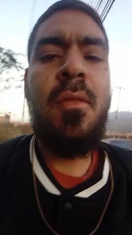 A man with a beard and mustache is recording a selfie-style video outdoors. He is wearing a black jacket with white stripes on the collar and an orange shirt underneath. A gold chain is visible around his neck. The background shows a street, trees, and a slightly blurry view of mountains in the distance. The man says "Buenos dias, buenos dias, inviertan, inviertan."