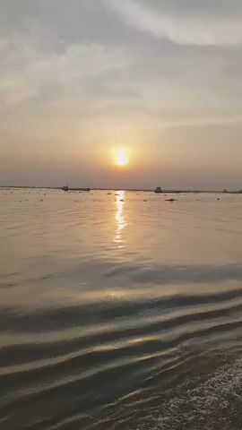 The video shows a wide expanse of water at sunset. The sun is low on the horizon, casting a bright reflection on the water's surface. There are some distant landmasses or boats visible on the horizon line. The water is filled with patches of vegetation or debris. The camera appears to be moving, creating ripples and waves in the water, with the wake from a boat in the foreground.