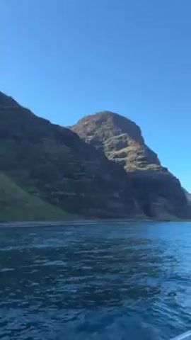 The video depicts a body of water with dark, rocky, and verdant coastal cliffs rising from its edge. The water's surface is textured with small waves and a deep blue color. The sky is clear and a lighter blue, with the sun obscured behind the peaks of the cliffs, casting a soft light on the scene. The perspective appears to be from a moving vessel, indicated by the movement of the water. There are no people visible.