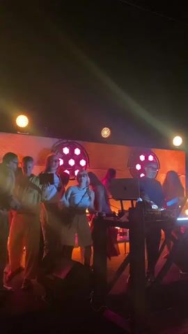 A group of people are on a stage with colored lights and a DJ booth. A DJ is behind the DJ booth with a laptop. One person is holding a tablet and looking at it. Another person is holding a phone and recording. The stage is lit with purple and yellow lights.