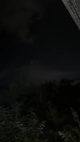 The video shows a dark outdoor scene at night. There is a lot of vegetation, including trees and bushes. The sky is dark with some faint clouds visible. The camera pans across the scene, providing a view of the landscape.
