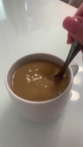 In this close-up shot, a person's hand with pink-painted fingernails is stirring a light brown liquid in a white ceramic mug. The mug sits on a reflective, smooth white surface, possibly a table. The liquid appears to be coffee or tea with milk or cream, as indicated by its color and the swirling patterns created by the spoon. The spoon itself is metallic and appears to have an ornate design on its handle. The lighting suggests an indoor setting, possibly during the daytime given the bright reflections on the table. No text is visible in the frame. The scene is simple and focuses on the act of preparing a drink.
