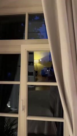 The video shows a room interior through a window. A large floor lamp with a white shade and a wooden tripod base is in the center of the frame, casting a bright yellow light on the wall behind it. To the right of the lamp, a blue sofa is visible, partially covered in plastic wrap. On the left side of the frame, a bookshelf is seen. The window itself is divided into panes by white mullions. A sheer white curtain is drawn to the right, partially obscuring the view. The room appears to be dimly lit, with the lamp being the primary light source. The reflection of the room on the window glass is also visible, showing the furniture and the lamp. The overall impression is of a cozy, interior space viewed from the outside.