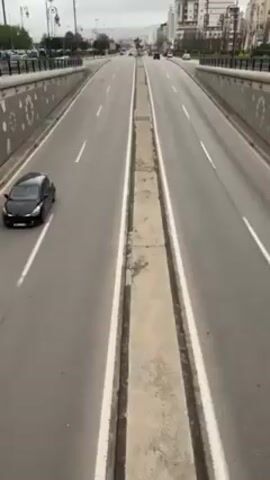 The video shows a view from a bridge overlooking a wide, straight city street with multiple lanes in each direction, divided by a concrete median. Cars are driving on both sides of the street, although the traffic appears light. The overpass is decorated with a pattern of circles and stars. Buildings are visible in the background on either side of the street. The sky is overcast.