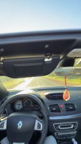 The video shows a view from the interior of a car driving down a straight, paved road on a sunny day. The car's top is down, revealing a clear blue sky with the sun shining brightly. The road is flanked by green fields with what appear to be olive trees enclosed by wire fences. Power lines run parallel to the road. The car's dashboard and steering wheel are visible in the foreground, with a decorative pendant hanging from the rearview mirror. Another car is parked on the side of the road in the distance.