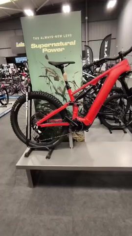 The video shows a red electric mountain bike on display in a bicycle shop. The bike is positioned on a stand and is the primary focus of the camera's attention. Behind the bike, a large green banner is visible with text that reads "THE ALWAYS-NEW LEVO" and "Supernatural Power." The background reveals other bicycles and store displays, indicating a retail environment. The camera moves slightly, offering different perspectives of the bike, and at one point, a price tag is visible hanging from the handlebars, indicating the bike's cost and available financing options. The lighting suggests an indoor setting, possibly under fluorescent lights. No people are directly interacting with the bike in the frame.