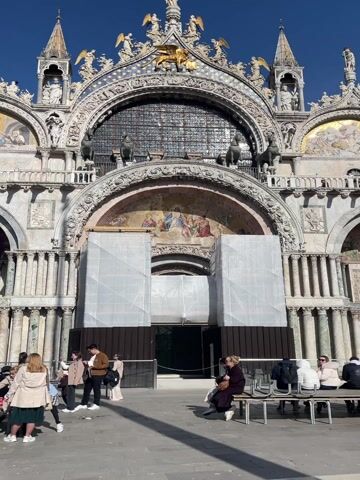 The video captures a bright, sunny daytime scene in St. Mark's Square in Venice, Italy. The prominent subject is the ornate facade of St. Mark's Basilica, a grand architectural masterpiece featuring multiple arches, intricate mosaics depicting religious scenes, and numerous sculptures, including a large golden winged lion and the famous bronze horses above the central upper arch. The basilica's roofline is adorned with spires and statues.

In the foreground, a considerable number of people are gathered in the square. Many are seated on benches scattered throughout the plaza, while others are standing, walking, and observing the surroundings. The central lower arch of the basilica's facade is partially covered by white scaffolding or tarps, indicating ongoing maintenance or restoration work.

The sky is clear and blue with only a few wispy clouds. Pigeons can be seen on the ground among the people. The overall atmosphere appears relaxed, with individuals enjoying the pleasant weather and the iconic Venetian landmark.