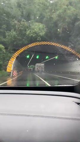 The video begins by showing a view from inside a car, looking out through the windshield at a tunnel entrance. The windshield is wet with raindrops, and there are water streaks on the glass. Yellow and black striped markings are visible on the tunnel archway, along with orange and white checkered barriers on the left side. Inside the tunnel, green lights are illuminated, and there are several vehicles ahead, including a truck with a trailer. The road surface appears wet and reflective, with more green lights visible further into the tunnel. The camera pans slightly as the car enters the tunnel. The interior of the car is dimly lit, with a portion of the dashboard visible in the foreground. The video continues to show the view from inside the car as it drives through the tunnel, with the wet windshield and interior dashboard remaining visible. The green lights and the truck ahead continue to be the main focal points through the windshield. The video then cuts off.
