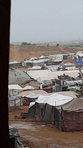 The video shows a high-angle, obstructed view of a refugee camp. The camp consists of many tents, predominantly white and dark gray, clustered close together on a barren, muddy landscape. In the background, a small hill or embankment is visible. The sky is overcast, creating a dull, grey light. The video is shot through a frame, possibly a window, partially obscuring the view. The overall atmosphere is bleak and gives the impression of crowded, impoverished living conditions.