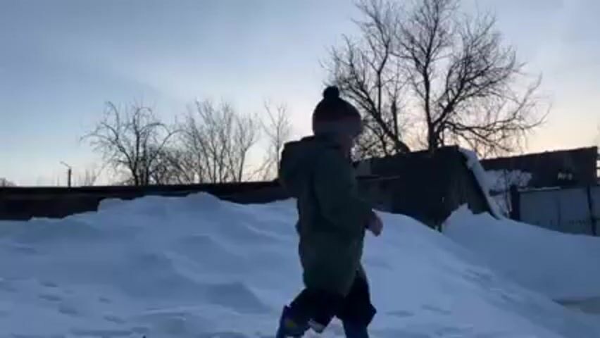 A young child in a winter coat and hat walks away from the camera on a snowy path. The child is wearing a dark green winter coat, a knit hat with a pompom, and blue boots. The path is covered in snow, with deeper drifts to the side. In the background are several trees with bare branches, a small building with a faded exterior, and a fence. The sky is overcast with a hint of sunset glow. The light is soft and diffused, creating long shadows. The camera follows the child as they walk along the path.