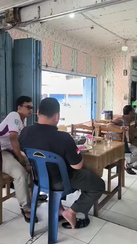 The video shows the interior of what appears to be a restaurant or food stall. The walls are decorated with a patterned wallpaper featuring pastel pink, green, and white stripes. Fluorescent lights are mounted on the ceiling. Several tables with brown tablecloths are set up, with chairs around them. In the foreground, a man wearing a black shirt and dark pants sits at a table with his back to the camera, facing away from the viewer. A blue plastic chair is visible. To his right, another man in a plaid shirt is seated, looking towards another table. Further back, a man in a brown shirt is sitting at a table. To his right, a man wearing a black cap and a black shirt is seated. There is a punching bag hanging in the background near a doorway. Several other people are visible in the background, some seated at tables. On the tables, there are plates, glasses, and bottles. The floor is tiled with a white and pink geometric pattern. The scene appears to be a casual gathering or mealtime.