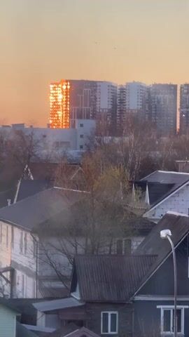 The video captures a sunset over an urban or suburban landscape in Akulovo (Gp Kubinka), Russia. The scene primarily features buildings and trees under a warm, orange sky. In the foreground, there are older, smaller houses with dark, sloped roofs, some appearing to have solar panels. A street light is visible on the right. In the midground, leafless trees and some with budding leaves stand between the houses and larger structures.

The main subject is a prominent, modern high-rise building in the background, whose windows are brilliantly reflecting the intense orange light of the setting sun. The building appears to be part of a larger complex of similar modern residential buildings. The camera zooms in on this reflective building, causing the reflected sunlight to become a vibrant, abstract, circular bokeh effect. The overall impression is one of a serene evening with striking natural light interacting with architectural elements. No people or text are visible.