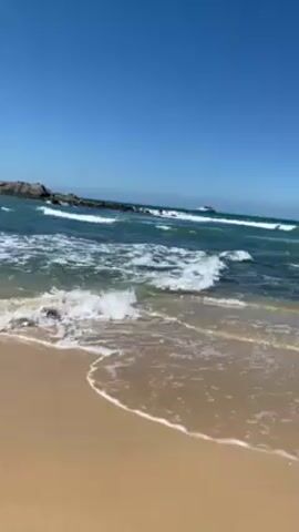 The video shows a sandy beach with gentle waves rolling onto the shore. The sand is light brown in color. The water is a mix of turquoise and white, with the white representing the foam of the waves. In the distance, there's a rocky outcropping to the left, and a small boat or ship can be seen further out on the water. The sky is a clear, bright blue. The camera appears to be handheld, with slight movements. The overall scene conveys a calm, sunny day at the beach.