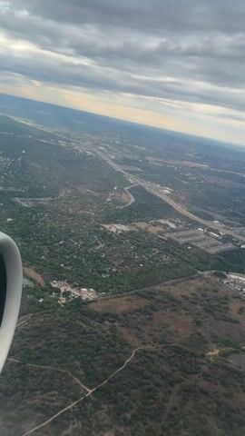 The view from an airplane shows an expansive landscape below. There is a mix of residential areas with dense tree coverage, a highway network with multiple lanes and off-ramps, and open fields. There is also a small, dark pond. The sky is overcast with a mix of gray and some light, suggesting partial sunlight. Part of the airplane engine is visible in the lower left corner of the frame. The scene is shot at a diagonal angle looking down.