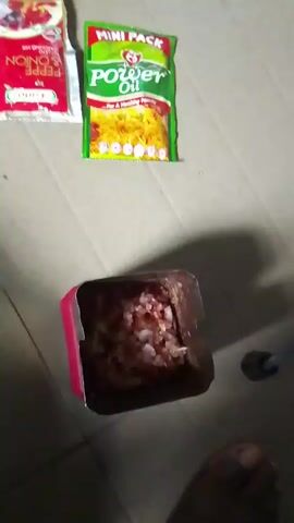 A close-up, top-down shot shows chopped raw meat and onions in a pink and silver container. To the left of the container are two small packets, one red and one green, with text and images on them. The green packet reads "MINI PACK Power Oil" and depicts a bowl of noodles. The red packet has text that is mostly obscured but appears to include "PEPPER & ONION SEASONING MIX". A person's foot is visible at the bottom of the frame. The scene takes place on a floor with a tiled or wood-like pattern.