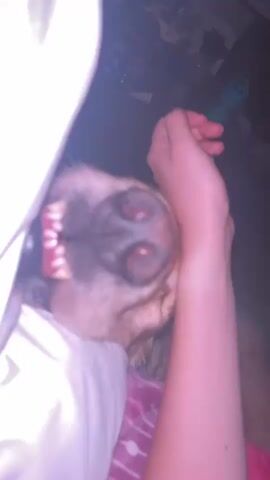 A dog's snout is close-up, with its mouth open to reveal its teeth. A person's arm is visible, with their hand stroking the dog's head. The dog appears to be lying down on a bed with white and pink bedding. The lighting is dim.