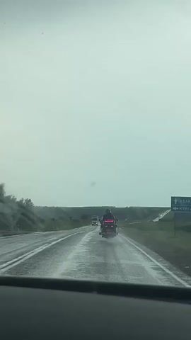 The video captures a view from inside a vehicle driving on a wet, asphalt highway under an overcast, grey sky. The road is reflecting the gloomy atmosphere, indicating it is likely raining or has recently rained. In the distance, a motorcycle with its brake lights illuminated is traveling ahead, followed by a large truck. The surrounding landscape appears to be rural with rolling hills and vegetation. The scene suggests a journey on a highway, possibly in a rural or semi-rural area of Kurgany, Russia, during daylight hours. The weather conditions create a somber or subdued mood. There is no visible text in the video.