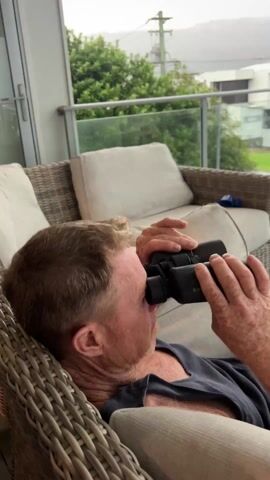 A man with fair hair and a grey top lounges on a wicker sofa on a covered patio. He is looking through binoculars. He lowers the binoculars and the camera pans to show a view of a neighborhood of houses and the ocean in the distance. It is overcast and raining.