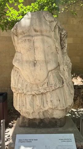 This media file shows a large, weathered stone sculpture of a torso, likely of a ruler, on display outdoors. The sculpture is heavily damaged, with the head and limbs missing, but the detail of the musculature of the abdomen and the belted garment is still visible. The sculpture is resting on a grey concrete base, which is itself on a darker stone foundation.

The background features lush green foliage from an overhanging tree casting dappled sunlight onto the sculpture. Behind the sculpture and tree, a modern brick wall creates a distinct contrast with the ancient artifact. To the left, a dark trash bin and a metal post are partially visible. The scene is bathed in bright daylight, suggesting it is mid-day, and the weather appears to be clear.

At the base of the sculpture, a white plaque with text in both English and Hebrew is mounted. The English text reads: "Statue of a Ruler, from the Temple of Augustus in Samaria. Acquired by Harvard University. Only discovered in the Harvard excavation at Samaria." The Hebrew text is a translation of the English. The overall impression is that of an archaeological artifact preserved and presented for public viewing in an outdoor setting. The location is identified as al-Eizariya, Palestinian Territories.