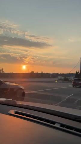The video captures a sunset view from inside a car. The camera is positioned behind the dashboard, looking out through the windshield. The sun is low in the sky, casting a warm orange glow across the clouds and the landscape. Trees and a highway are visible in the distance, with other vehicles on the road. The scene appears to be on a highway or an elevated road, as evidenced by the barrier and the bridge structure that comes into view later in the video. The mood is serene and picturesque, typical of a sunset drive.