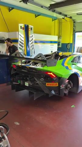Inside a garage painted with a yellow top and red floor, a lime green and yellow racing car, without rear tires, dominates the scene. The car features a large rear wing, a complex carbon fiber rear diffuser, and Pirelli tire branding. A man stands to the left, leaning against a work surface and engrossed in a phone call. Another man enters the frame and stands near the back of the car. Various pieces of equipment are scattered around the garage, including a blue and silver cabinet, air hoses, and tools, emphasizing the workshop setting.