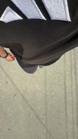 The media file shows a first-person perspective of someone walking on a paved surface, likely a sidewalk or street. The subject is wearing dark clothing, including pants and a black t-shirt or sweatshirt with a prominent white logo on it. The logo appears to be an Adidas logo with three stripes forming a triangle. The person is also wearing a watch on their left wrist. The ground is a textured gray concrete. Sunlight casts shadows, indicating it is daytime. The overall scene is mundane, depicting a simple act of walking outdoors. There are no other people visible in the frame. The location is identified as Taraz, Kazakhstan, but this context is not visually apparent from the image itself.