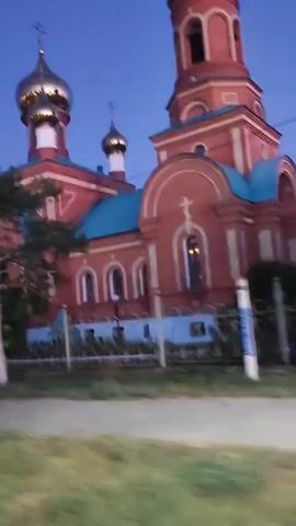 The video shows a red brick church with gold domes and blue accents. It is dusk, and the sky is a deep blue. The church is partially obscured by a tree in the foreground. The camera is moving, suggesting it is mounted on a vehicle. There is a fence in front of the church. The overall scene is peaceful and serene.