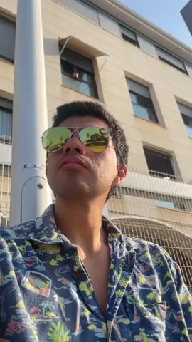 A low-angle, close-up shot focuses on a man wearing aviator sunglasses with mirrored lenses and a brightly colored Hawaiian shirt. The man is looking upwards, his mouth slightly open. Behind him, a multi-story building with numerous windows is visible. The building appears to be made of beige-colored material. In the background, a white pole is positioned to the left of the man, and a metal fence is visible to the right. The lighting suggests it is daytime, with sunlight illuminating the scene from the side, casting shadows. Reflections of a street scene, possibly with palm trees, can be seen in the man's sunglasses.