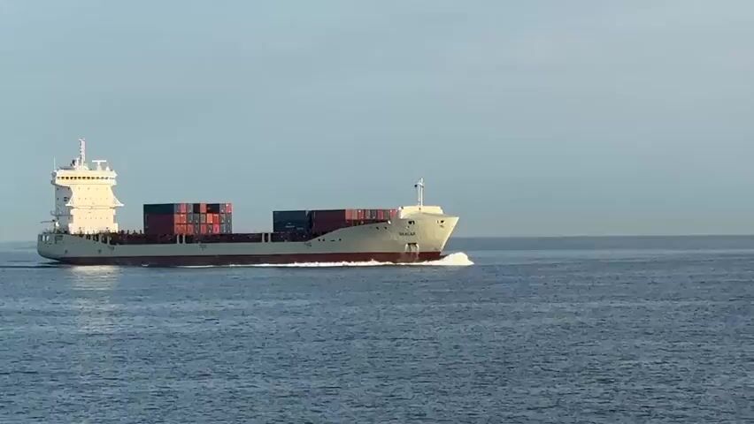 This media file depicts a large cargo ship sailing across a calm, blue sea under a clear, pale blue sky. The ship, which appears to be a container ship, is painted white with a red waterline. It is laden with numerous stacked cargo containers, some of which are red and some are dark blue. The ship has a tall, multi-story superstructure at the stern, with windows visible. A wake of white foam trails behind the ship, indicating it is in motion.

The time of day appears to be either early morning or late afternoon, given the soft, even lighting and the absence of strong shadows. The weather seems pleasant and clear, with no clouds visible in the sky. The scene is one of serene maritime activity, with the vast expanse of the ocean surrounding the vessel. The ship's name, "SEALAN," is visible in white lettering on its bow. The overall mood is peaceful and tranquil, suggesting a smooth journey across the water. There are no people clearly visible in the frame, focusing the attention on the ship and the sea. The location is unknown, though a note suggests a context related to Germany.