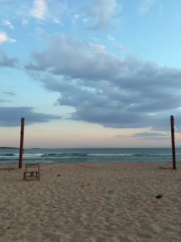 This media file captures a tranquil scene on a sandy beach in Voula, Greece, likely during sunset or sunrise given the soft, warm light and pastel hues on the horizon. The sky is a blend of clear light blue at the top, transitioning to paler blue and hints of pink closer to the horizon, dotted with scattered white and grey clouds.

In the foreground, a wide expanse of light brown sand stretches towards the calm sea. Two tall, weathered wooden poles stand upright on the sand, one on the left and one on the right, typically used to support a beach volleyball net, which is absent in this frame. To the left of the center, a small, rustic wooden chair with a woven seat rests on the sand, facing the ocean. A small, dark object, possibly a piece of litter or a shoe, is visible on the sand near the right pole.

The sea exhibits gentle waves rolling towards the shore, with the water appearing a clear greenish-blue. The horizon line is visible, with a faint, distant landmass on the far left. No people are present, lending the scene an air of peaceful solitude. There is no visible text in the image.