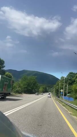 A car is driving on a two-lane road. The road has a yellow line on the right side and white lines in the middle. The car is in the right lane. There are mountains in the background and trees on the sides of the road. There are also other cars on the road. The sky is partly cloudy.