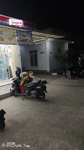 At night, outside a convenience store or gas station, two motorcycles are parked on a concrete lot. One motorcycle is in the foreground, facing left, with a person sitting on it wearing a light-colored headscarf and red pants. The motorcycle is loaded with bags. In the background, another motorcycle is parked to the right, with three people standing and sitting near it. The building behind them is illuminated by artificial lights. A sign with images of vehicles is visible above the building's entrance.