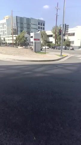 The video shows a view from a street looking across an intersection towards a modern office building complex. The street appears to be paved and dark. There is a small empty lot on the near side of the intersection, surrounded by a short fence. A sign is visible on the lot, possibly indicating the building or complex. A white Jeep-like vehicle is parked on the far side of the intersection. The building complex is multi-storied, with a mix of glass and white facade. A traffic light is visible at the intersection. The sky is blue with sparse clouds. A siren sound is heard in the background.